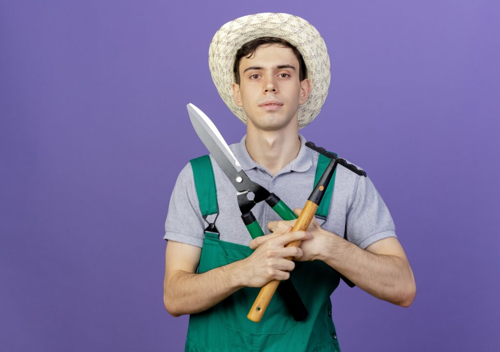 confident-young-male-gardener-wearing-gardening-hat-holds-rake-clippers-min